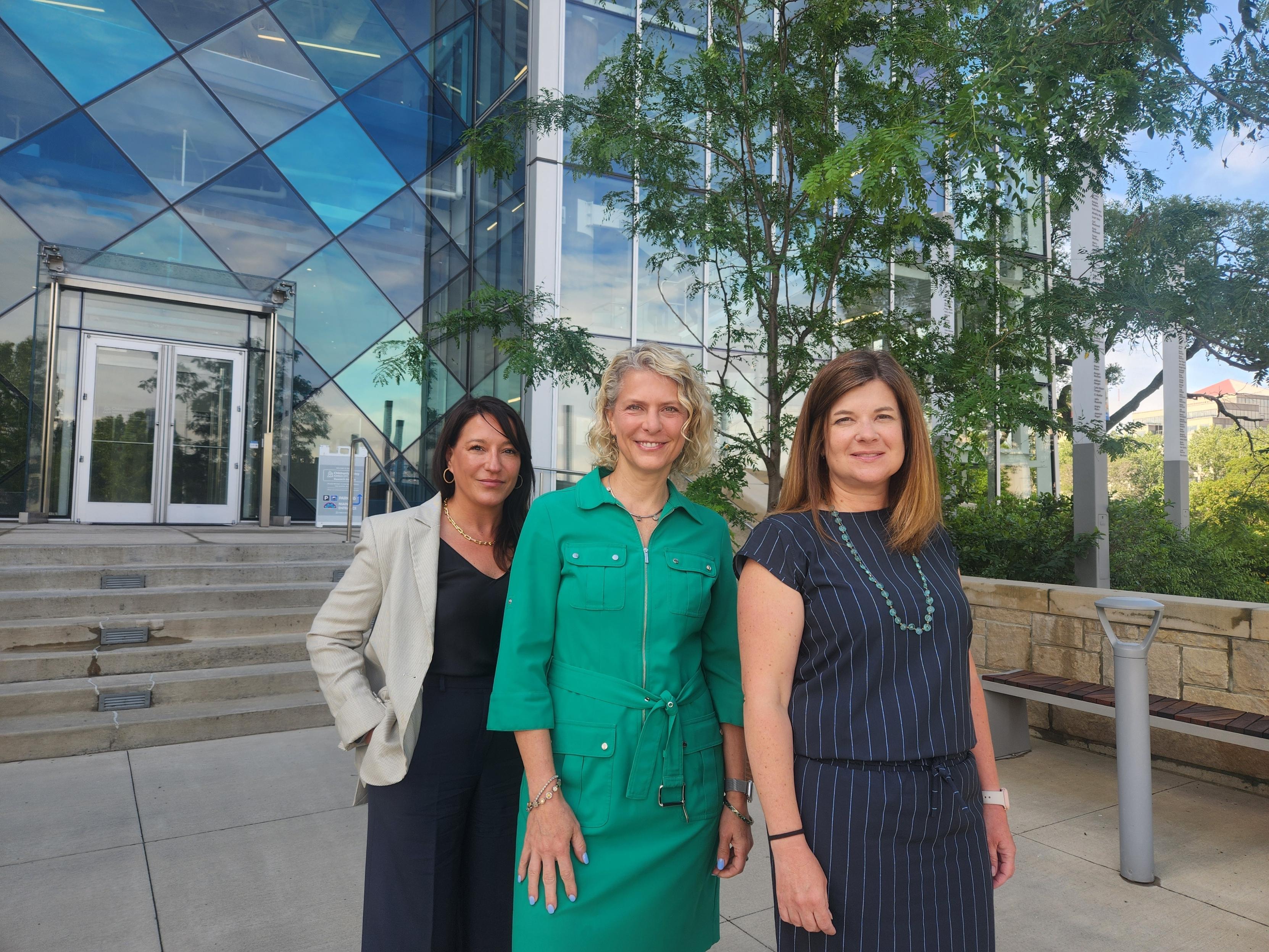 Three women, part of the Children’s Mercy Clinical Genetics and Genomics Lab, pose in front of a building with a facade of glass tiles in shades of blue. 